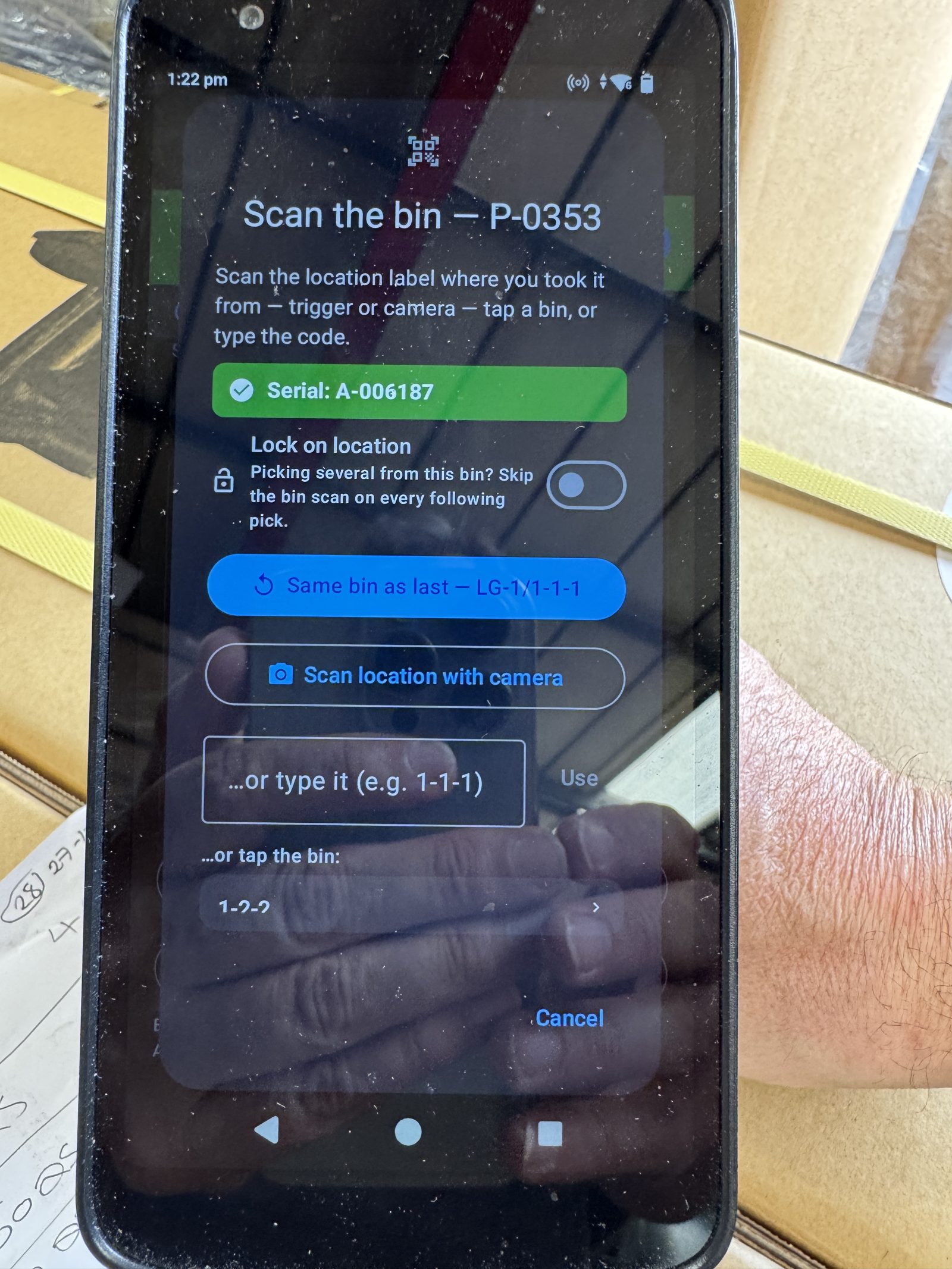 A warehouse operator holding a Zebra handheld mid-scan, prompted to scan the bin they took stock P-0353 from, standing among stacked cardboard boxes
